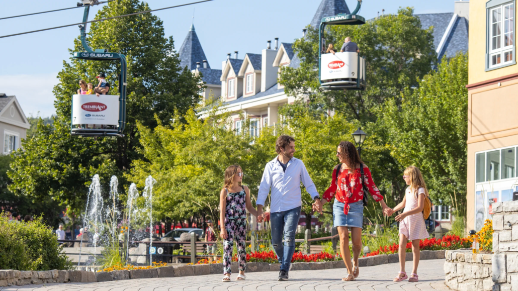 Summer family activity vacation packages at Les Suites Tremblant, showing a family strolling past scenic courtyard fountains and trees with panoramic gondolas overhead in Mont-Tremblant, Quebec.